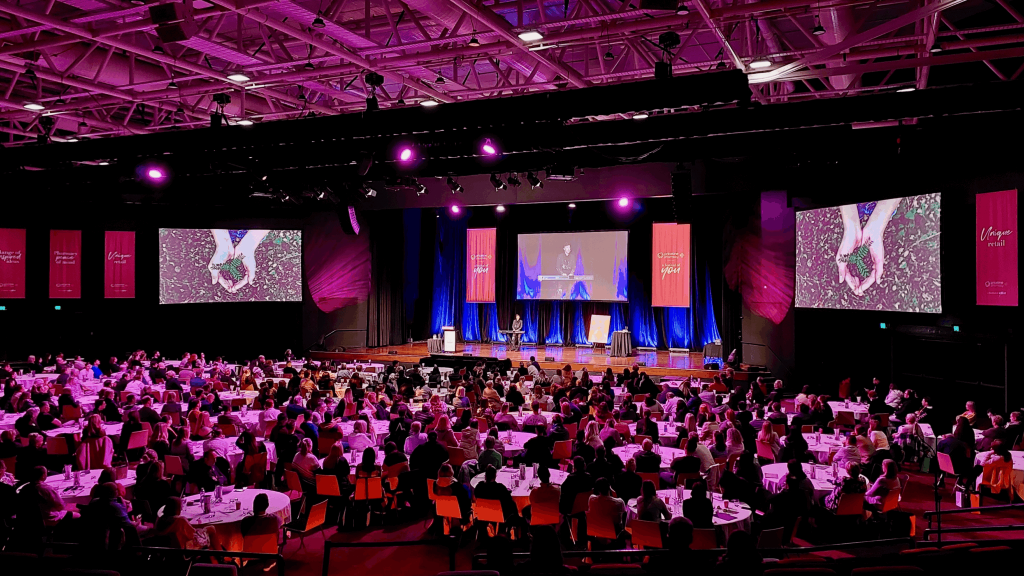 Sarah Rowan speaking on stage at a large conference with painting displayed on screens