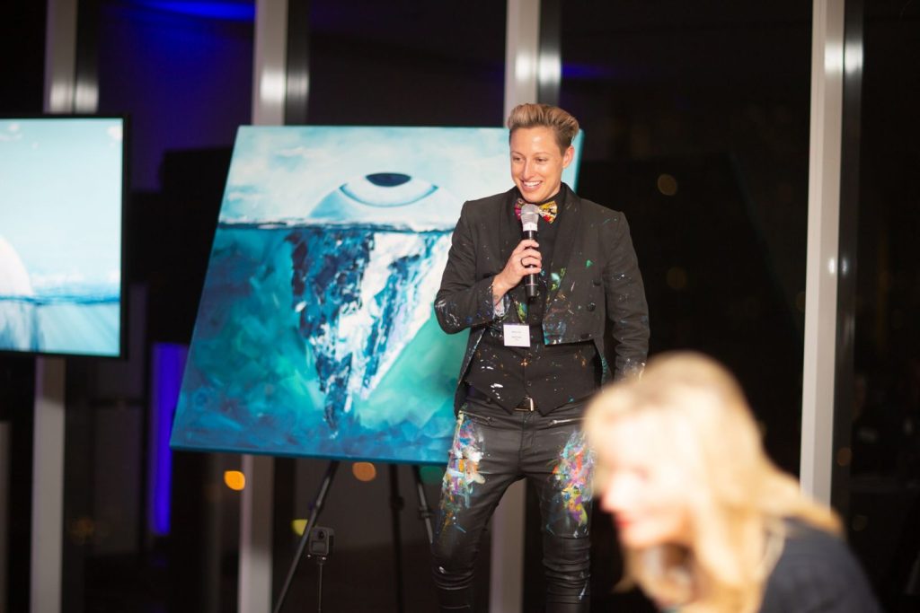 Sarah Rowan speaking on stage beside her iceberg painting