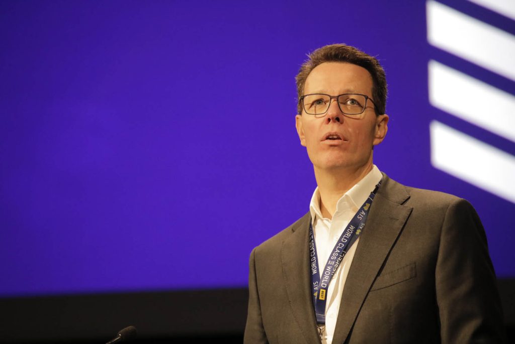 Kieren Perkins speaking on stage in front of a purple screen