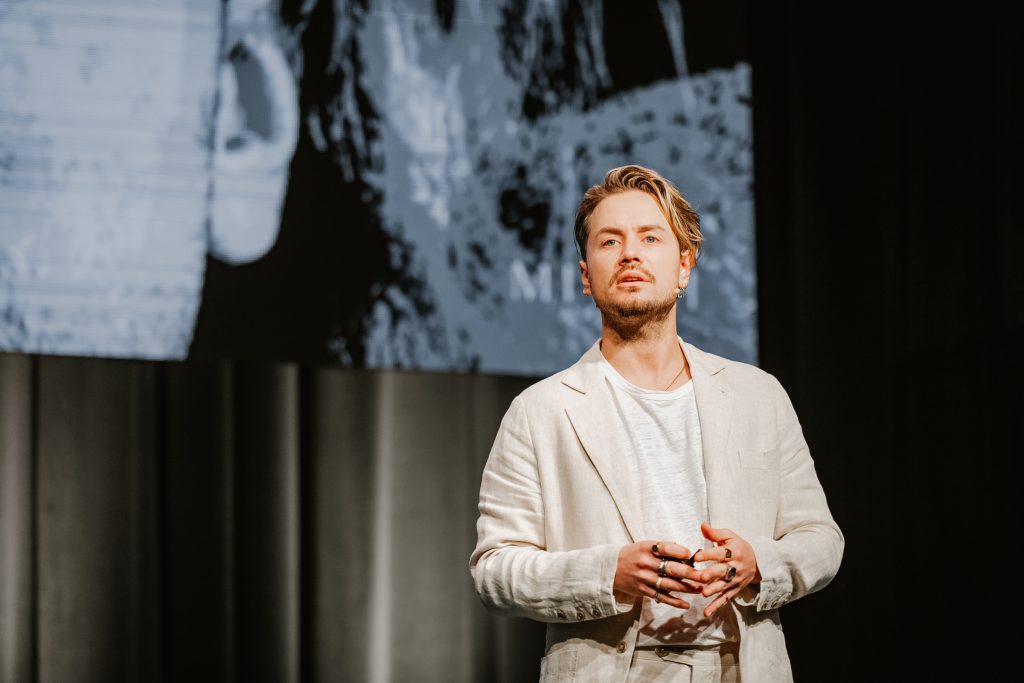 Mitch Wallis speaking on stage in a cream suit
