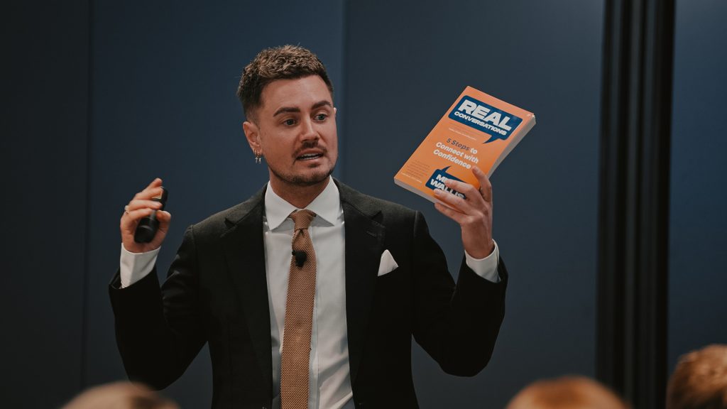 Mitch Wallis holding his book Real Conversations on stage
